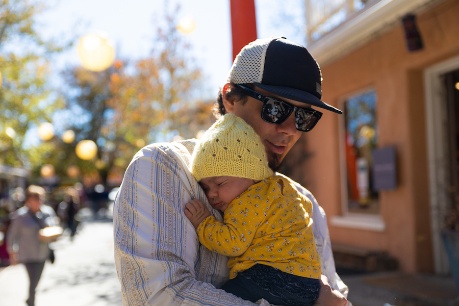 Father in hat & sunglasses holding baby in yellow outfit.