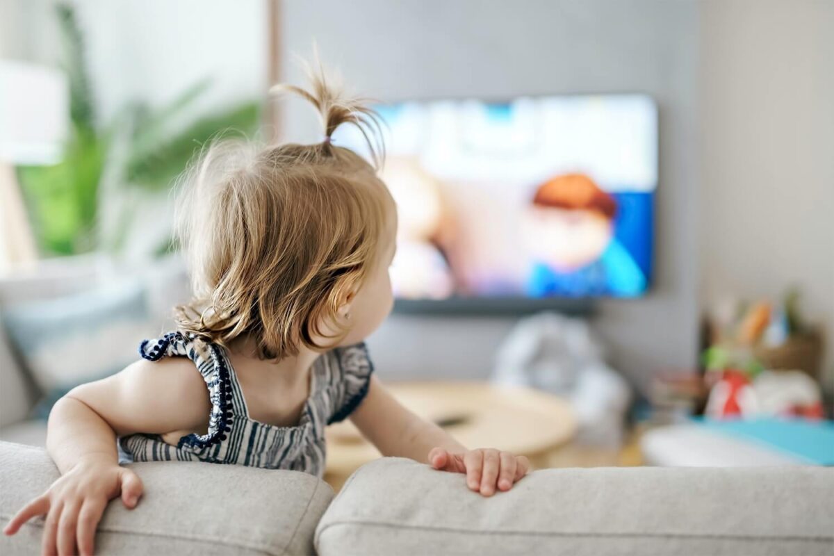 Toddler climbing on a couch looking backward to see TV.